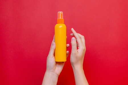 Female hand holding a orange spray cosmetic bottle on the solid red background.