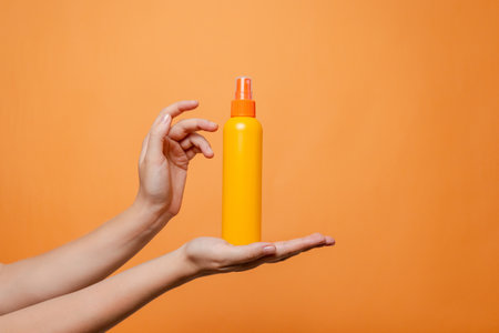 Hand holding a orange spray cosmetic bottle on the solid orange background.
