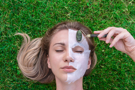 Woman's face with cosmetic face mask and with roller smoothing wrinkles in the grass. Spa, healthy,...