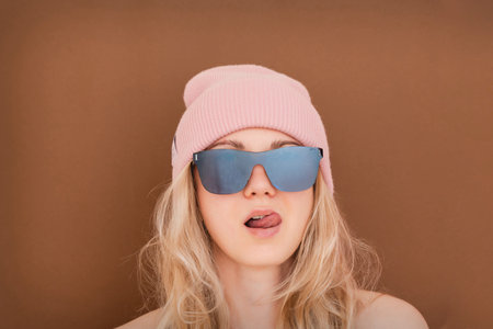 Young woman posing with pink beanie and mirror sunglasses, sticking out her tongue