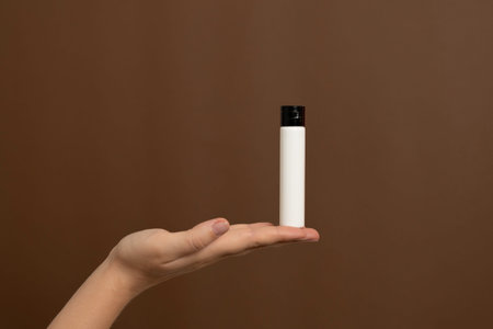 Hand presenting a blank white cosmetic bottle against a brown backgroundの写真素材