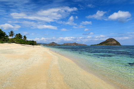 White sand clear beach with palm trees and mountains on cloudy blue sky background at Sumbawa island, Indonesiaの写真素材