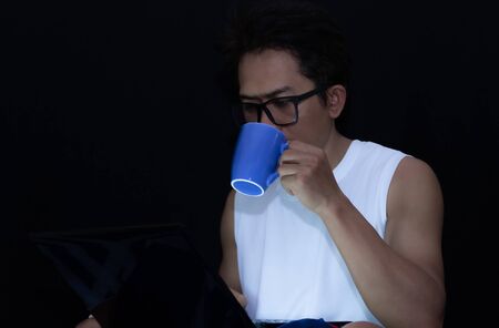 Low light view and copy space. A man wear glasses drink coffee while using a laptop. He works online...
