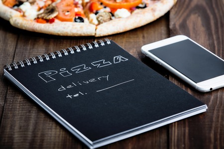 Close-up of pizza, a mobile phone and a notebook with the text: Pizza delivery. background wooden table. Notebook black with white textの写真素材