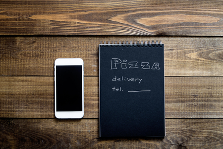 Close-up of mobile phone and notebook with the text: Pizza delivery. background wooden table. Notebook black with white textの写真素材