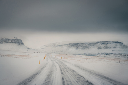 Winter landscape of Iceland's road during snowstorm with the snow-capped mountain as a background and frozen road as a foreground.の写真素材