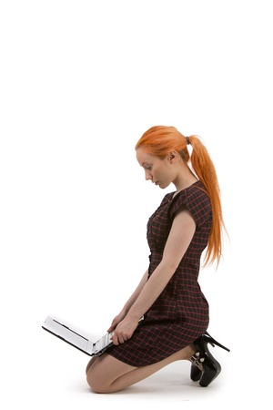 Side view of an attractive young woman with her long red hair tied in a ponytail kneeling on the floor in a stylish black dress and high heels reading her laptopの写真素材