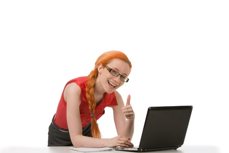 Smiling young redhead woman in glasses sitting at her laptop giving a thumbs up gesture of success isolated on whiteの写真素材