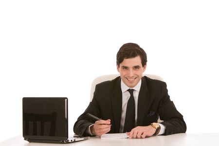 Smiling handsome young businessman sitting writing at his desk with an open laptop in front of himの写真素材