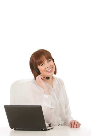 Friendly attractive young female call centre operator sitting at her desk with a laptop talking to a client on her headset and smiling at the viewerの写真素材