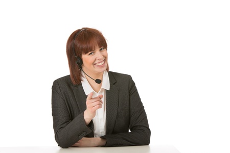 Smiling attractive young female call centre operator wearing a headset pointing at the viewer while seated at her desk, on a white backgroundの写真素材