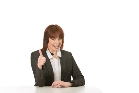 Happy woman in a headset sitting at a table with a lovely cheerful smile giving a thumbs up of approval and success, isolated on whiteの写真素材