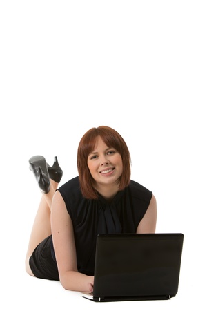 Relaxed woman with a sweet smile wearing a black dress and high heels lying on her stomach using her laptop on the floor, on a white backgroundの写真素材