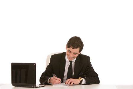 Smiling handsome young businessman seated at his desk in front of his laptop writing notes on a notepadの写真素材