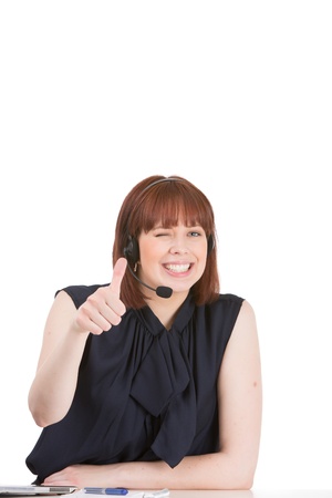 Playful attractive young woman wearing a headset smiling and giving a thumbs up of agreement or approval while winking at the camera, on whiteの写真素材