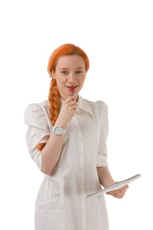 Smiling thoughtful young redhead woman standing holding a notebook with her pen to her mouth isolated on whiteの写真素材
