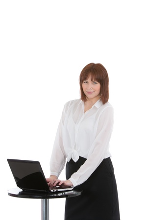 Beautiful young business woman standing typing on her laptop at a small round metal table , three quarter portrait isolated on whiteの写真素材