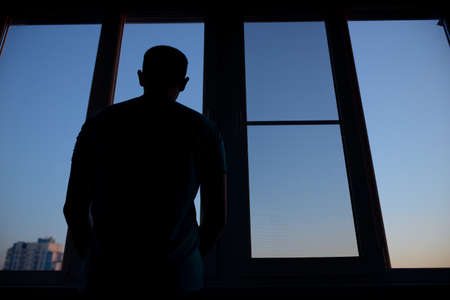 a man looks out the window at sunset from the balcony view from the back. self-isolation in an apartment during a pandemicの写真素材