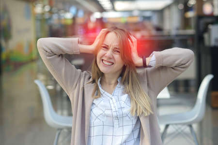 Young woman suffering from headache in a shopping center cafe. Portrait of frustrated young woman with headache and frowning while from noiseの写真素材