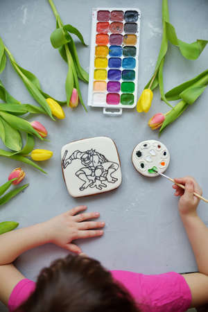 a girl paints a gingerbread with food dyes. A coloring book for children lies on a gray table. the...