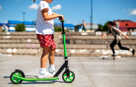 preschool girl riding a scooter in red shorts and a white T-shirt on the playground on an early sunny hot summer morning. active rest in the park.の写真素材