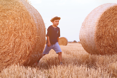 Young cute country boy in has walking happily through wheat field near hay stack or bale. Active outdoors on the beach.の写真素材