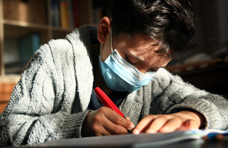 Teenager boy wearing face mask doing homework at home and writing notes.