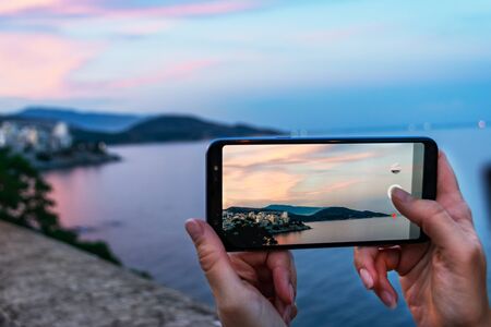 Female hands holding smartphone taking picture of city with beautiful sunset sky.  Sightseing concept.の写真素材