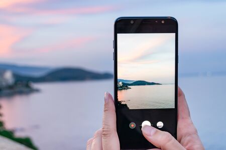 Female hands holding smartphone taking picture of city with beautiful sunset sky.  Sightseing concept.の写真素材