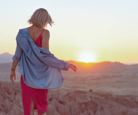 Back view of the young woman with blue shirt and pink dress standing at edge of the cliff and enjoying beautiful view of valley during sunsetの写真素材