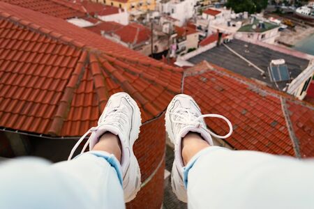 Woman sitting on the edge of building and her legs with white sneakers dangling against red rooftopsの写真素材