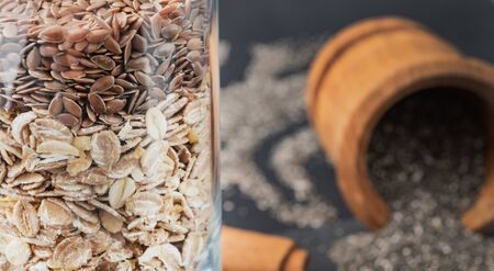 Close up oats and flaxseeds in a glass with unfocused wooden bowl of chia seeds on background. Paleo...