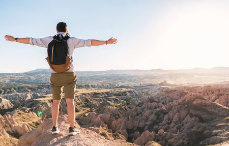 Caucasian man tourist with backpack and spread hands standing on the edge of cliff, enjoying sunset on canyons and valleys of Cappadocia.の写真素材
