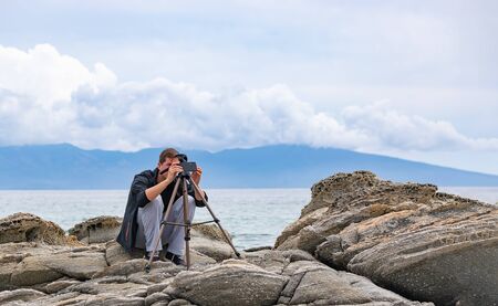 Young handsome male photographer sitting on rocks by the sea setting up mobile phone tripod....