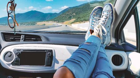 Female legs in ripped jeans and blue sneakers inside car with mountains landscape at background. Woman relaxing during road tripの写真素材