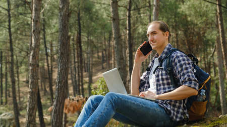 Young man in forest talking on smartphone and working on laptopの写真素材