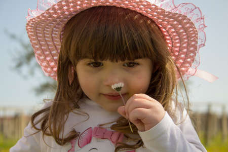 Portrait of an adorable little girl in a pink hat sniffing a daisy sitting in an outdoor field.