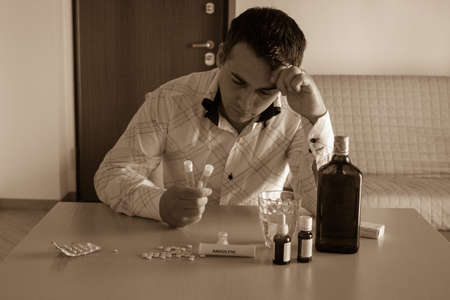 Worried man sitting and leaning on the table at home with anxiolytics and antidepressants in front while holding a test tube in anus. Side effects of psychiatric drugs on male ejaculationの写真素材