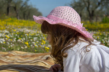 Image of an adorable little girl in a pink hat lying on a blanket in the middle of a field of flowers during a vacation.の写真素材