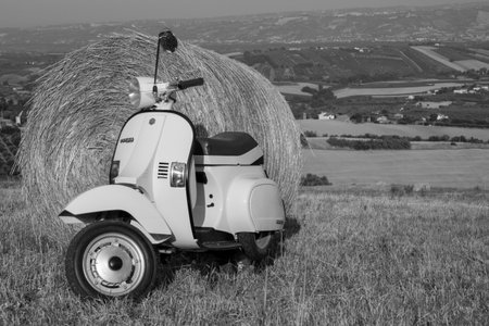 Image of an old Piaggio vespa motorcycle parked in a field with straw bales and stunning view in the background. Black and white photos. Dates 29-06-2022 Florence Italyのeditorial素材