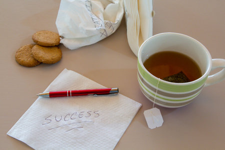 Image of a napkin with success written on it, and cookies in the background, a cup of tea, a pen and more napkins. Reference to ambitions and ideas during breakfast.の写真素材