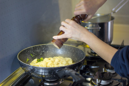 Image of a woman's hands seasoning freshly cooked pasta in a pan with a pepper mill. Italian...