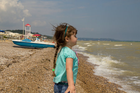 Image of an adorable little brunette girl playing at the seaside throwing stones into the water. Beach vacation trip to Italyの写真素材
