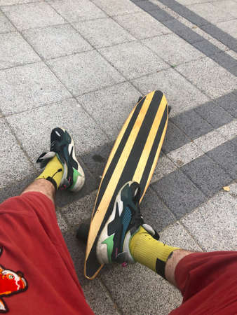 An anonymous man in red shorts sitting on the pavement near his longboard after skating along the embankment. Skate made of wood and sandpaper, painted yellow. Fun active hobby outdoors activity.の写真素材