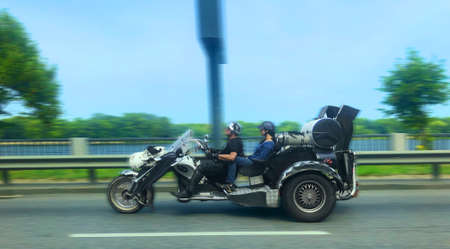 Family couple of man and woman riding on big beautiful old-fashioned three wheel motorcycle wearing protective road helmet. Blurred trees and billboard in background, highway in front. Kyiv, Ukraine.の写真素材