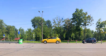 A yellow new car is driving along the road, standing out in the general traffic. A park with green trees, road signs, and a dividing strip with posts In the background. Driving school concept.の写真素材