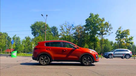 A red new car is driving along the road, standing out in the general traffic. A park with green...