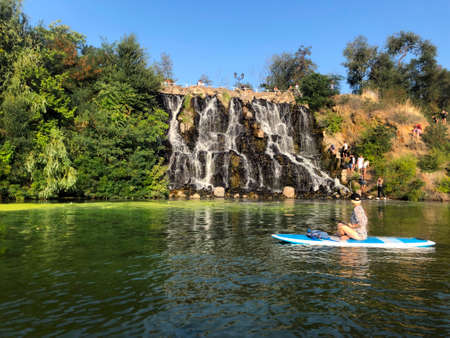 Monastyrsky Island, Dnipro, Ukraine. A group of tourists descends to artificial waterfall. A girl on sup rafting down the river. The waterfall is surrounded by trees against a blue sky without cloudsのeditorial素材