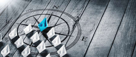 Blue Paper Boat Leading A Fleet Of Small White Boats With Compass Icon On Wooden Table With Sunlight...