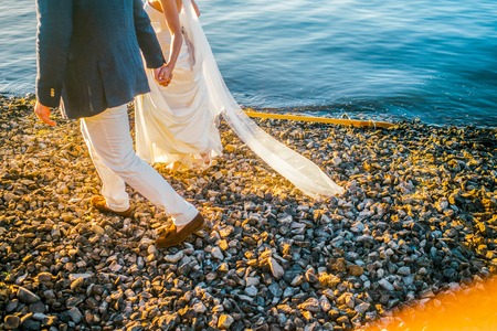 Wedding stroll along the beach with stones and yellow sunlights  by bride in white dress with and groom in blue jacket, white pants and brown shoesの写真素材